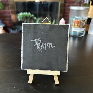 May include: A square black tile with a white handwritten signature and date on the back, featuring a gold-colored triangular metal hanging hook at the top. The tile is displayed upright on a small, light-colored wooden tripod easel stand.