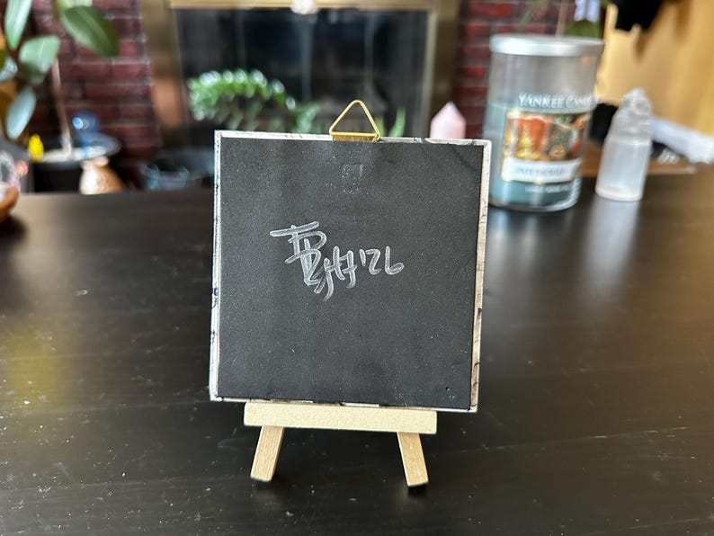 May include: A square black tile with a white handwritten signature and date on the back, featuring a gold-colored triangular metal hanging hook at the top. The tile is displayed upright on a small, light-colored wooden tripod easel stand.