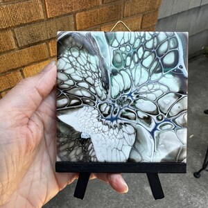 May include: A square, abstract art tile with a black wooden easel. The tile features a fluid design in shades of white, black, blue, and green. The artwork is displayed on a small, black easel, perfect for tabletop display.