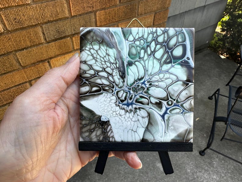 May include: A square, abstract art tile with a black wooden easel. The tile features a fluid design in shades of white, black, blue, and green. The artwork is displayed on a small, black easel, perfect for tabletop display.