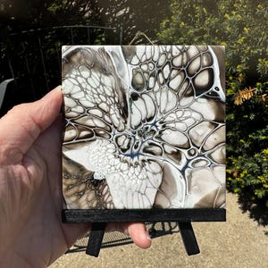 May include: Square abstract art tile with a black wooden easel. The tile features a fluid art design in shades of white, gray, and black, with intricate patterns resembling cells or veins. The artwork is displayed on a small black easel.