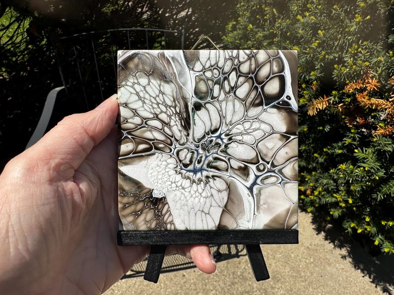 May include: Square abstract art tile with a black wooden easel. The tile features a fluid art design in shades of white, gray, and black, with intricate patterns resembling cells or veins. The artwork is displayed on a small black easel.