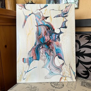 May include: Abstract acrylic painting on canvas. The artwork features a cream-colored background with flowing patterns in shades of blue, pink, and black. The design has a fluid, organic appearance with a cracked, textured effect.