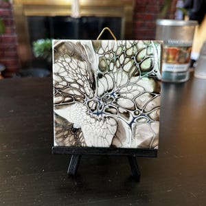 May include: Square ceramic tile with an abstract design in shades of white, black, and gray, displayed on a small black easel. The artwork features a fluid, cell-like pattern with hints of blue.