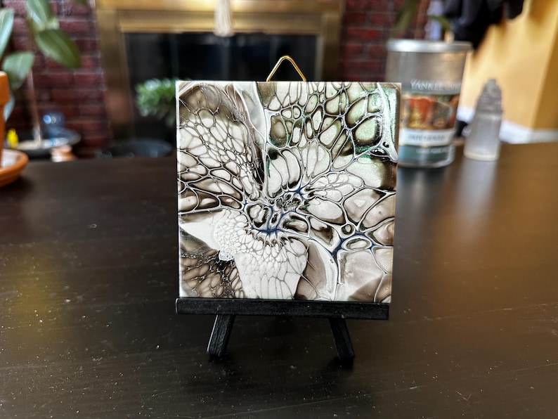 May include: Square ceramic tile with an abstract design in shades of white, black, and gray, displayed on a small black easel. The artwork features a fluid, cell-like pattern with hints of blue.