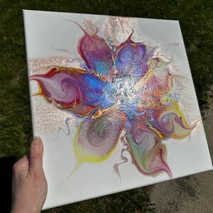 Pu&ograve; includere: Una tela quadrata con un dipinto astratto realizzato con la tecnica acrilica su sfondo bianco. Il design centrale mostra un motivo floreale vorticoso nei toni del blu iridescente, magenta, giallo e vernice color oro metallizzato.