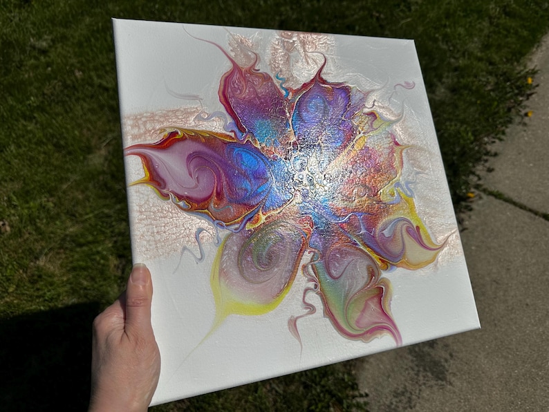 Pu&ograve; includere: Una tela quadrata con un dipinto astratto realizzato con la tecnica acrilica su sfondo bianco. Il design centrale mostra un motivo floreale vorticoso nei toni del blu iridescente, magenta, giallo e vernice color oro metallizzato.