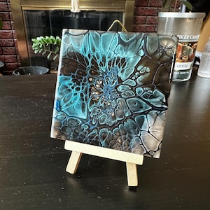May include: A square tile featuring an abstract acrylic pour design with swirling patterns of teal, black, and white. The tile is displayed on a small, light-colored wooden easel, showcasing a glossy finish and intricate cell-like structures.
