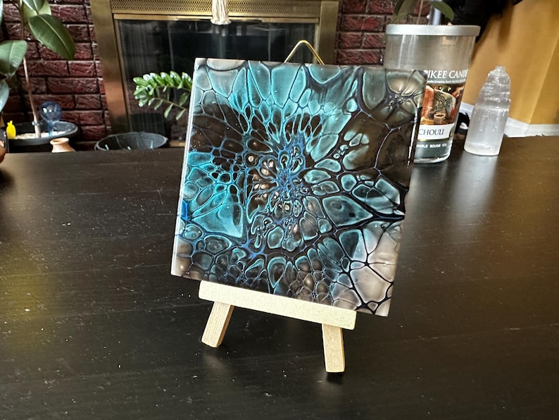 May include: A square tile featuring an abstract acrylic pour design with swirling patterns of teal, black, and white. The tile is displayed on a small, light-colored wooden easel, showcasing a glossy finish and intricate cell-like structures.