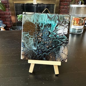 May include: A square tile featuring an abstract acrylic pour painting with a glossy finish. The design displays a central vortex pattern in shades of teal, black, and white, creating a web-like cell structure. The tile rests on a small wooden easel.
