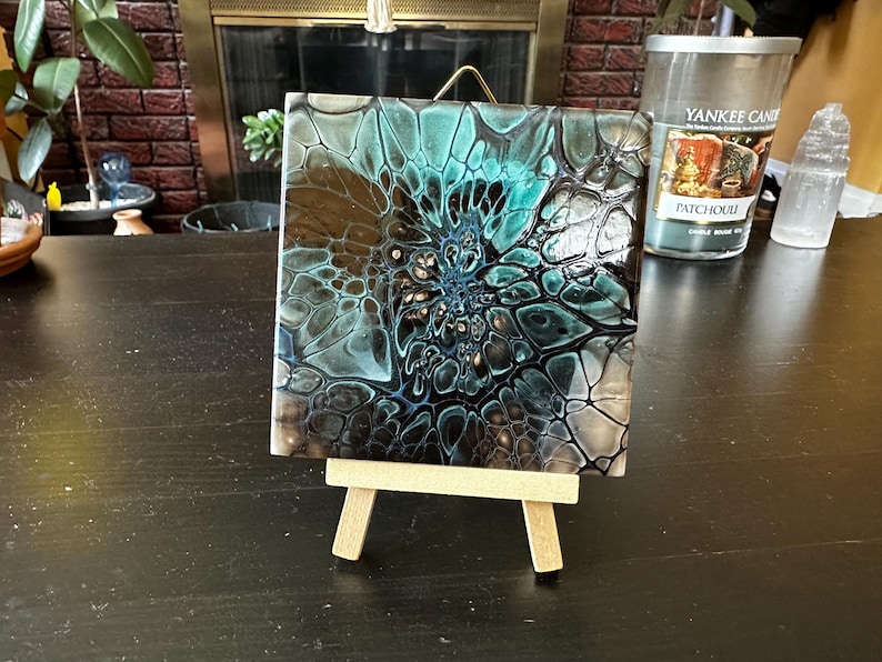 May include: A square tile featuring an abstract acrylic pour painting with a glossy finish. The design displays a central vortex pattern in shades of teal, black, and white, creating a web-like cell structure. The tile rests on a small wooden easel.
