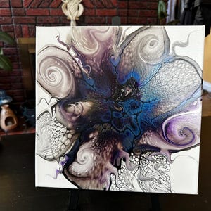 Puede incluir: Un lienzo cuadrado que presenta una pintura abstracta de vertido acr&iacute;lico con un estallido central de azul profundo y negro, rodeado de patrones arremolinados de plata met&aacute;lica, lavanda suave y blanco, creando efectos de celdas y encaje.
