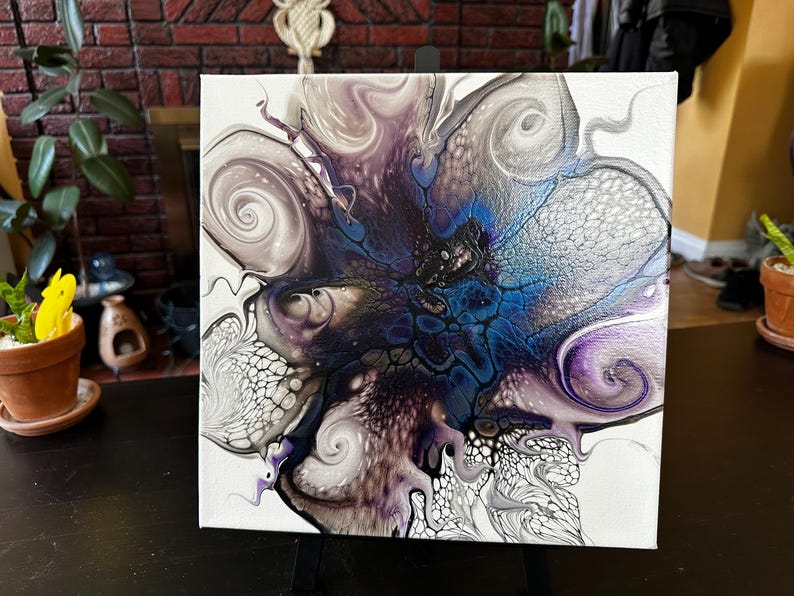 Puede incluir: Un lienzo cuadrado que presenta una pintura abstracta de vertido acr&iacute;lico con un estallido central de azul profundo y negro, rodeado de patrones arremolinados de plata met&aacute;lica, lavanda suave y blanco, creando efectos de celdas y encaje.