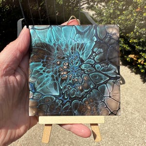 May include: A square tile featuring an abstract fluid acrylic pour design with metallic teal, black, and bronze colors. The pattern displays intricate, web-like cell structures radiating from the center. The tile is displayed on a small wooden easel.
