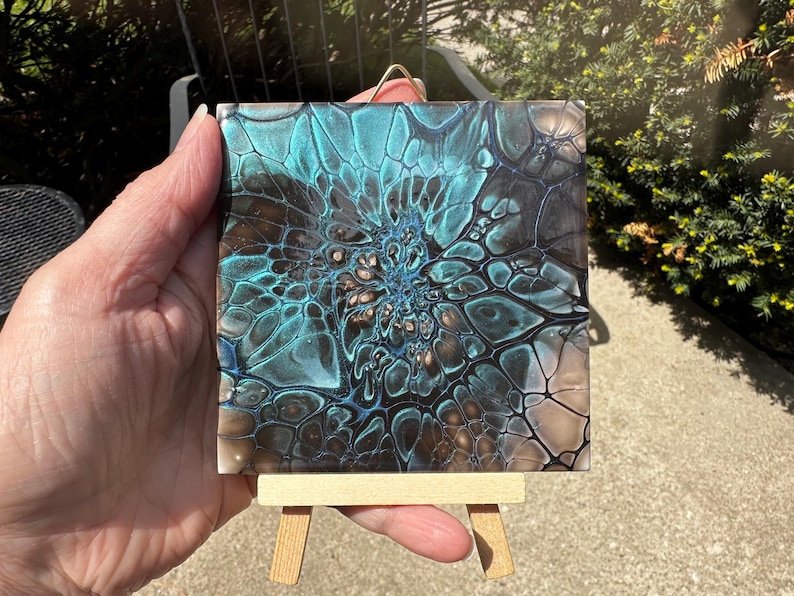 May include: A square tile featuring an abstract fluid acrylic pour design with metallic teal, black, and bronze colors. The pattern displays intricate, web-like cell structures radiating from the center. The tile is displayed on a small wooden easel.