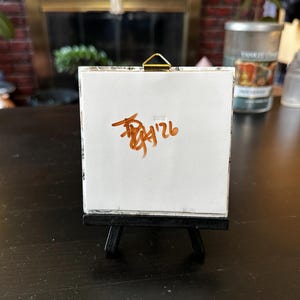May include: A small, white canvas on a miniature black easel. The canvas displays brown handwritten text, including the numbers "9'26". The easel is positioned on a dark wooden surface, with a blurred background featuring a candle and greenery.