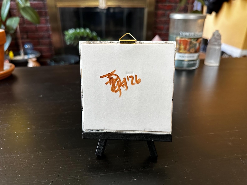 May include: A small, white canvas on a miniature black easel. The canvas displays brown handwritten text, including the numbers "9'26". The easel is positioned on a dark wooden surface, with a blurred background featuring a candle and greenery.