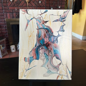 May include: Abstract acrylic painting on canvas. The artwork features a central, organic form with flowing lines in shades of blue, coral, and white, set against a cream-colored background. Gold accents add highlights to the piece.