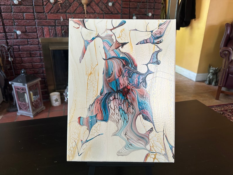 May include: Abstract acrylic painting on canvas. The artwork features a central, organic form with flowing lines in shades of blue, coral, and white, set against a cream-colored background. Gold accents add highlights to the piece.