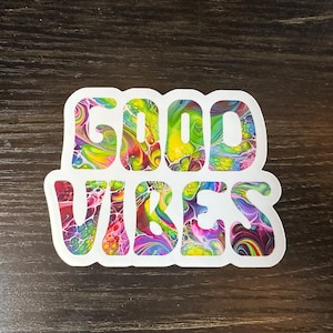 May include: A white sticker with the words "GOOD VIBES" in large, colorful, psychedelic-style lettering. The letters are filled with swirling patterns of red, yellow, green, blue, and purple. The sticker is on a dark brown wooden surface.