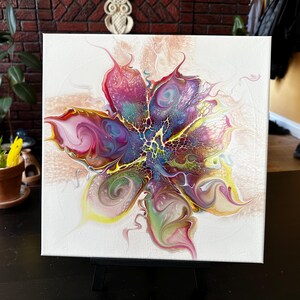 Pu&ograve; includere: Una tela quadrata che presenta un dipinto astratto a colata acrilica con un design centrale simile a un fiore. La composizione mostra vortici vibranti di magenta, viola, giallo e verde, accentuati da intricati motivi a merletto bianco.