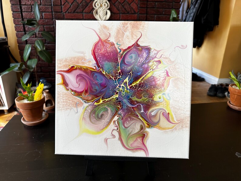 Pu&ograve; includere: Una tela quadrata che presenta un dipinto astratto a colata acrilica con un design centrale simile a un fiore. La composizione mostra vortici vibranti di magenta, viola, giallo e verde, accentuati da intricati motivi a merletto bianco.