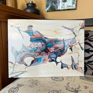 May include: Abstract acrylic painting on canvas. The artwork features a flowing design with shades of white, gold, blue, pink, and burgundy. The painting is displayed on a patterned surface.