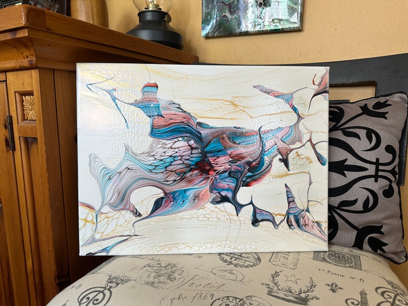 May include: Abstract acrylic painting on canvas. The artwork features a flowing design with shades of white, gold, blue, pink, and burgundy. The painting is displayed on a patterned surface.