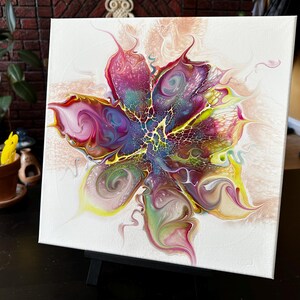 Pu&ograve; includere: Un dipinto su tela quadrata con un design floreale astratto creato con vernice acrilica fluida. La composizione mostra vortici vibranti di magenta, viola, giallo e verde, con intricati motivi a merletto che si irradiano dal centro.