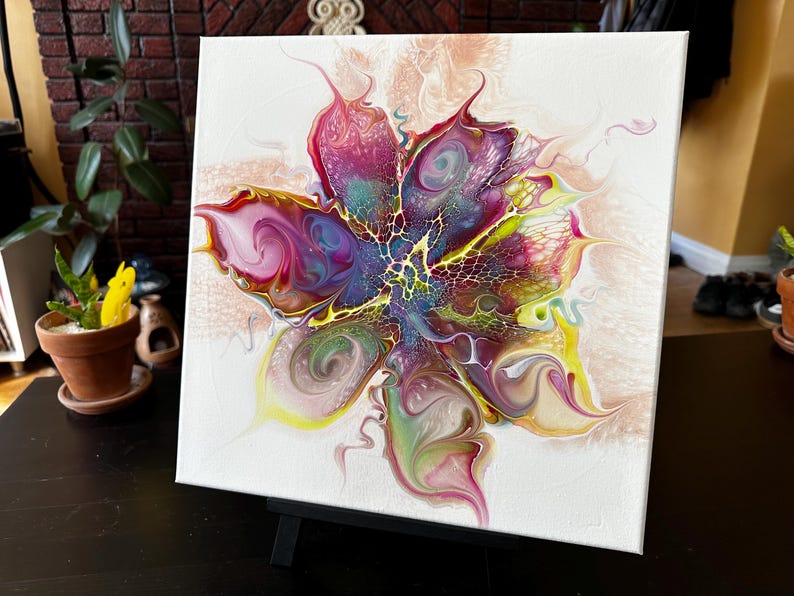 Pu&ograve; includere: Un dipinto su tela quadrata con un design floreale astratto creato con vernice acrilica fluida. La composizione mostra vortici vibranti di magenta, viola, giallo e verde, con intricati motivi a merletto che si irradiano dal centro.