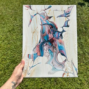 May include: Abstract acrylic painting on canvas. The artwork features a central design with flowing shapes in shades of blue, pink, and black against a white background. Gold accents add detail to the piece. The canvas is held by a person.