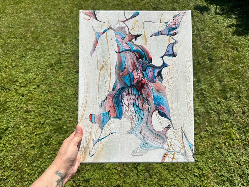 May include: Abstract acrylic painting on canvas. The artwork features a central design with flowing shapes in shades of blue, pink, and black against a white background. Gold accents add detail to the piece. The canvas is held by a person.