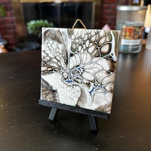 May include: Square ceramic tile with an abstract design in shades of white, black, and gray, displayed on a small black easel. The artwork features a fluid, cell-like pattern with hints of blue.