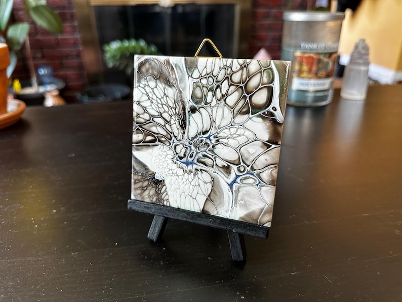 May include: Square ceramic tile with an abstract design in shades of white, black, and gray, displayed on a small black easel. The artwork features a fluid, cell-like pattern with hints of blue.