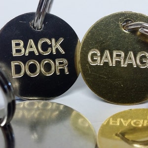 May include: Four round metal key tags with key rings. Two tags are gold with black lettering, one says "GARAGE" and the other says "BACK DOOR". The other two tags are silver with gold lettering.