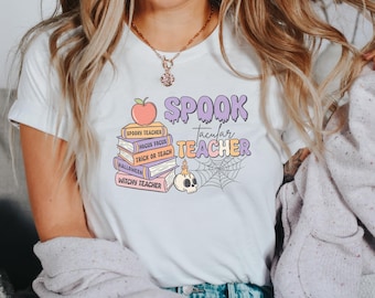 Könnte beinhalten: Ein weißes T-Shirt mit einer Grafik eines Bücherstapels mit den Aufschriften Spooky Teacher, Hocus Focus, Trick or Teach, Halloween und Witchy Teacher, gekrönt von einem Apfel, einem Totenkopf mit Kerze und einem Spinnennetz, sowie dem Text Spooktacular Teacher.