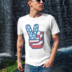 May include: White t-shirt featuring a peace sign design in red, white, and blue, with a distressed, vintage look. The peace sign incorporates the American flag's stars and stripes. The shirt is worn with blue jeans.