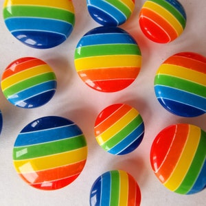 Puede incluir: Una colección de botones redondos y brillantes con rayas de arcoíris de varios tamaños. Los botones presentan un espectro vibrante de colores, incluyendo rojo, naranja, amarillo, verde, azul e índigo. Los botones están dispuestos sobre una superficie blanca.