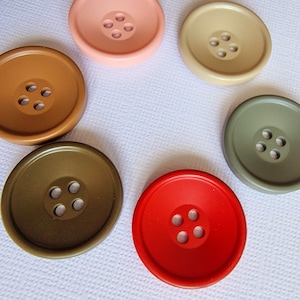 Puede incluir: Seis botones de diferentes colores con cuatro agujeros cada uno. Los botones están dispuestos en círculo sobre una superficie blanca. Los colores son rosa, marrón, beige, verde, rojo y verde oscuro.