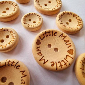 Puede incluir: Una colección de botones de madera con el texto "Little Man" y un símbolo de corazón grabado en cada botón. Los botones son de color marrón claro y tienen dos agujeros.