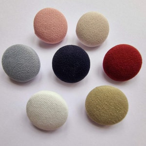 Könnte beinhalten: Sieben mit Stoff bezogene Knöpfe in verschiedenen Farben, darunter Rot, Blau, Beige, Rosa und Grau. Die Knöpfe sind rund und haben eine strukturierte Oberfläche.
