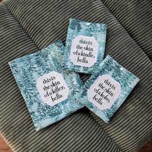 Puede incluir: Tres fundas rectangulares de tela con un estampado de bosque en color verde azulado y blanco. Cada funda tiene un cierre de broche blanco y un parche central blanco con texto en negro. Los parches dicen: this is the skin of a kindle, bella; this is the skin of a killer, bella; y this is the skin of a kobo, bella.