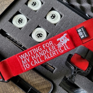 Könnte beinhalten: Ein schwarzer Koffer mit vier weißen Scheiben, jede mit einem schwarzen Logo. Ein roter Gurt mit weißem Text "WAITING FOR MY HANDLER TO CALL ALERT" und ein K9 Tactical Gear-Etikett sind ebenfalls sichtbar.