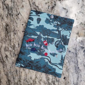 May include: A rectangular fabric pouch in shades of teal and dark blue featuring a printed landscape with a motorcycle, a red truck, and a cactus. The text "hoa hoa hoa hoa hoa" is embroidered in white across the center with a silver snap closure.