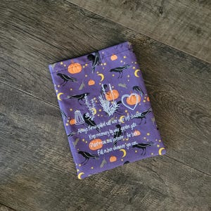 May include: A purple fabric pouch with a button closure. The pouch is decorated with illustrations of pumpkins, crows, moons, and text. The text reads: "Always throw spilled salt over your left shoulder..." and other phrases.