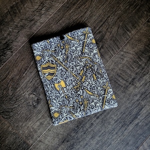 May include: A rectangular case with a black, white, and gold pattern featuring medieval weaponry and floral elements. The design includes swords, axes, shields, and decorative foliage. The case is set against a dark wood grain background.