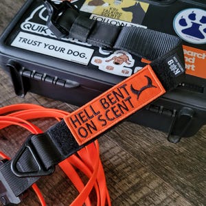 May include: Black tactical dog collar with an orange patch that reads "HELL BENT ON SCENT". The collar is next to an orange lead and a black case with stickers. The case has the text "TRUST YOUR DOG."