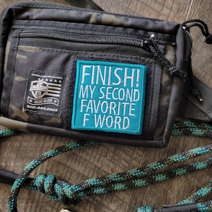 May include: Black and camouflage-patterned dog treat bag with a teal patch that reads "FINISH! MY SECOND FAVORITE F WORD." Includes a matching teal and black rope leash with a silver clasp. Made in the USA.