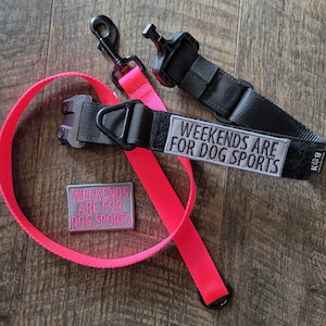 May include: A neon pink dog leash with a black metal clip and a black collar featuring a grey patch that reads WEEKENDS ARE FOR DOG SPORTS. A separate matching rectangular grey patch with pink embroidered text is included in the set.