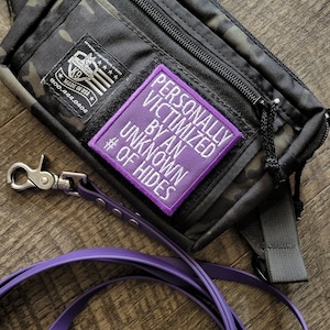 May include: A black and dark green camouflage fanny pack with a rectangular purple patch featuring the white text PERSONALLY VICTIMIZED BY AN UNKNOWN # OF HIDES. A purple leash with a silver metal clip is attached to the side of the bag.